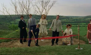 A groundbreaking ceremony in The Brutalist (2024) - A group of well-dressed 1950s figures stand in a rural landscape with bare trees, featuring a ceremonial red ribbon and shovel