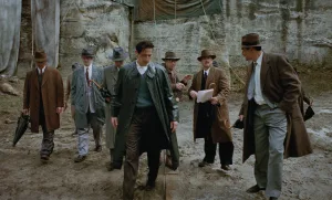 Construction site scene from The Brutalist (2024) - Architect in long green coat inspecting site with businessmen in 1950s attire against concrete wall backdrop