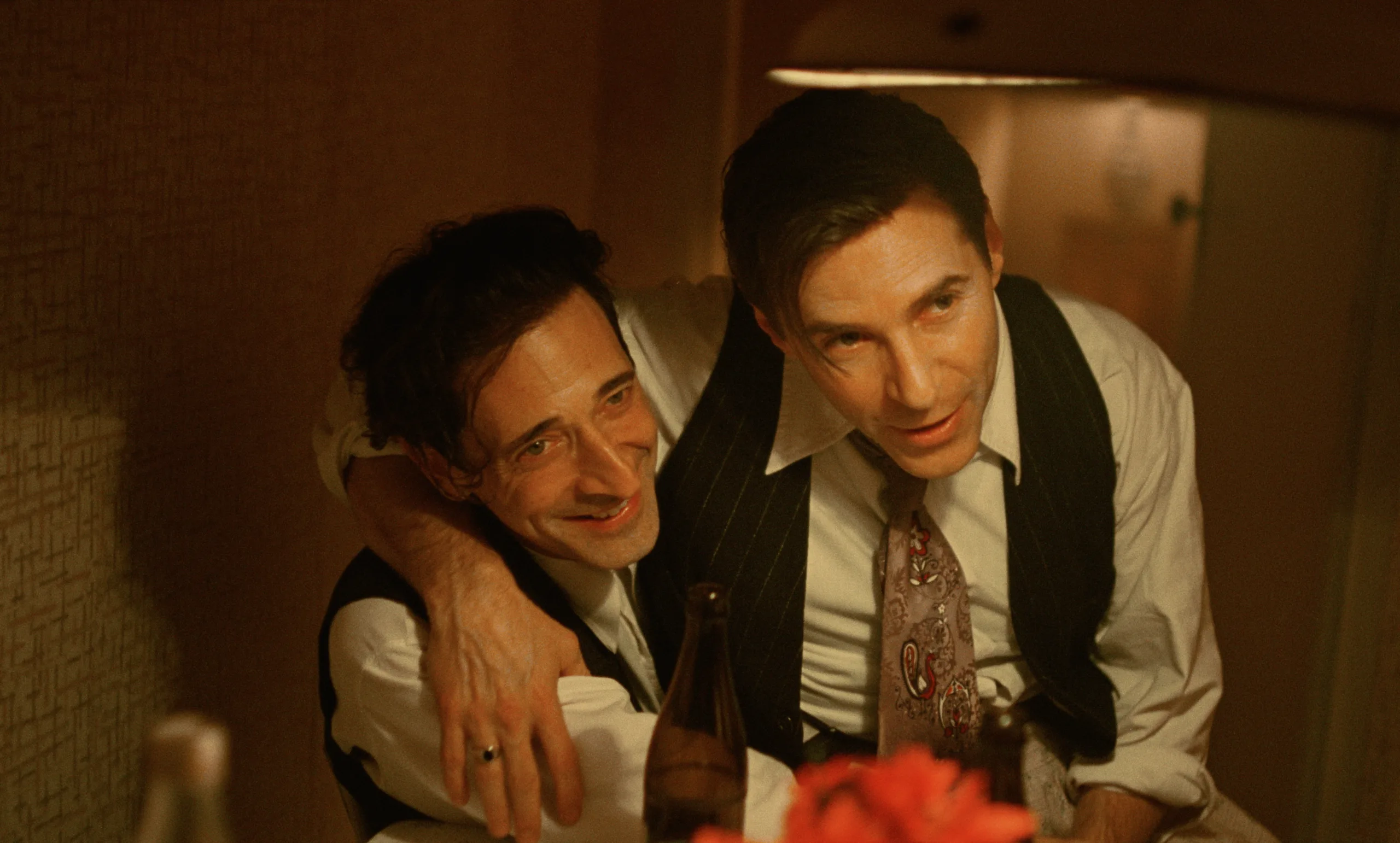 Adrien Brody and Guy Pearce in The Brutalist (2024) - A candid moment showing both actors in evening attire, sharing a warm interaction in warm, amber lighting