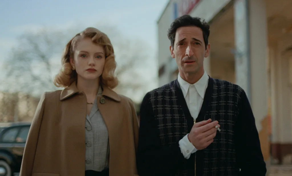 A scene from The Brutalist (2024) featuring Adrien Brody in a plaid cardigan and Felicity Jones in a camel coat, both in 1950s attire against a light background