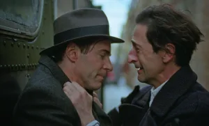 An intimate scene from The Brutalist (2024) showing a tense moment between two men, one wearing a period fedora hat, in close profile against a soft-focus background