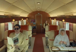 Benicio Del Toro os Zsa-Zsa Korda, Michael Cera os Bjorn kai i Mia Threapleton os Liesl se skini tis tainias The Phoenician Scheme mesa se aeroplano kathimenoi stis theseisme kokkino xali sto diadromo kai luxury aisthitiki